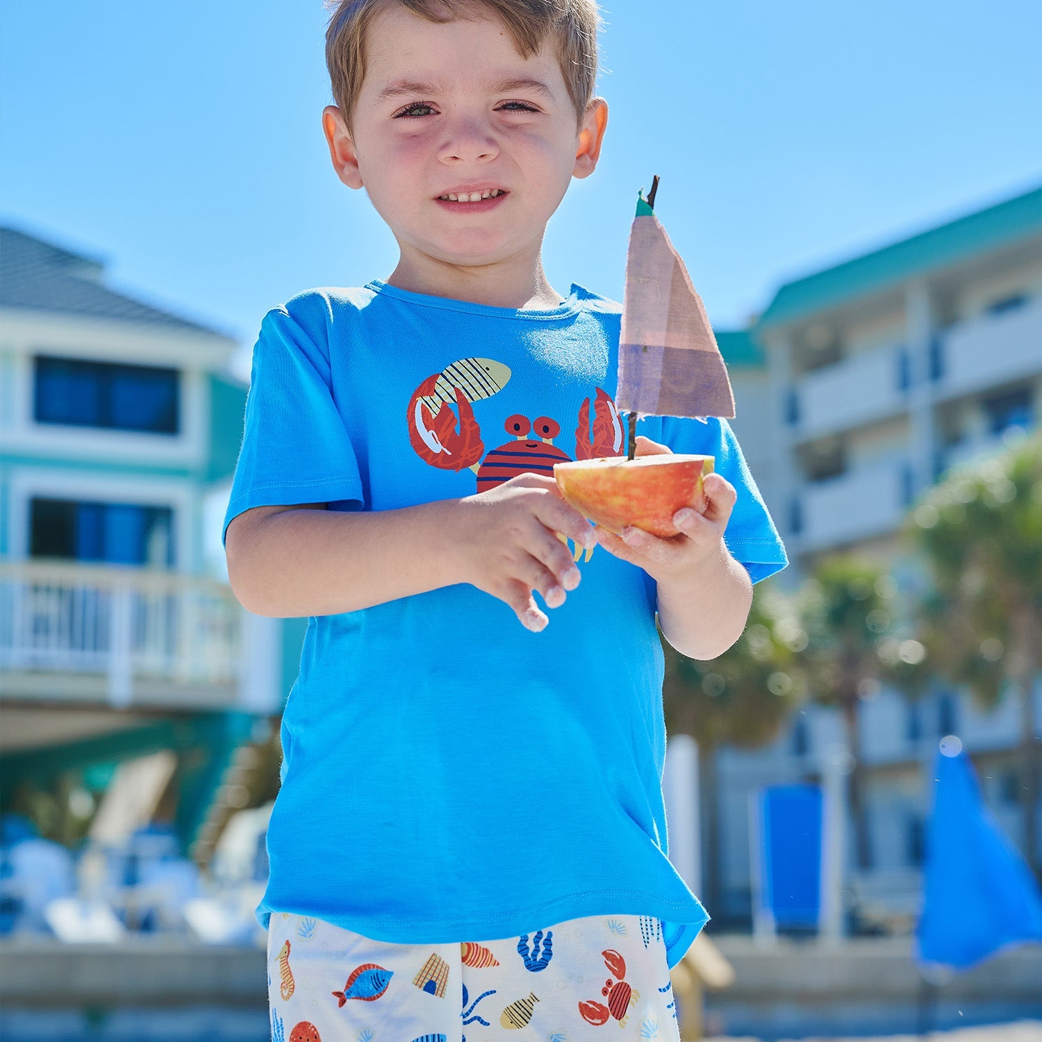 A young boy wearing the Magnetic Me Boys’ Deep Chic Magnetic Top & Short Set holds a small toy sailboat made from an apple in an outdoor setting with buildings and trees in the background.
