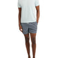 A man stands barefoot with hands in pockets against a white background, wearing the Onia Joey Crewneck Tee in light blue paired with patterned navy swim shorts.