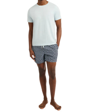 A man stands barefoot with hands in pockets against a white background, wearing the Onia Joey Crewneck Tee in light blue paired with patterned navy swim shorts.
