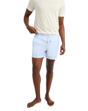A man stands barefoot against a plain background, wearing a white t-shirt and light blue Onia Charles 5" Swim Trunk made from quick-dry fabric.