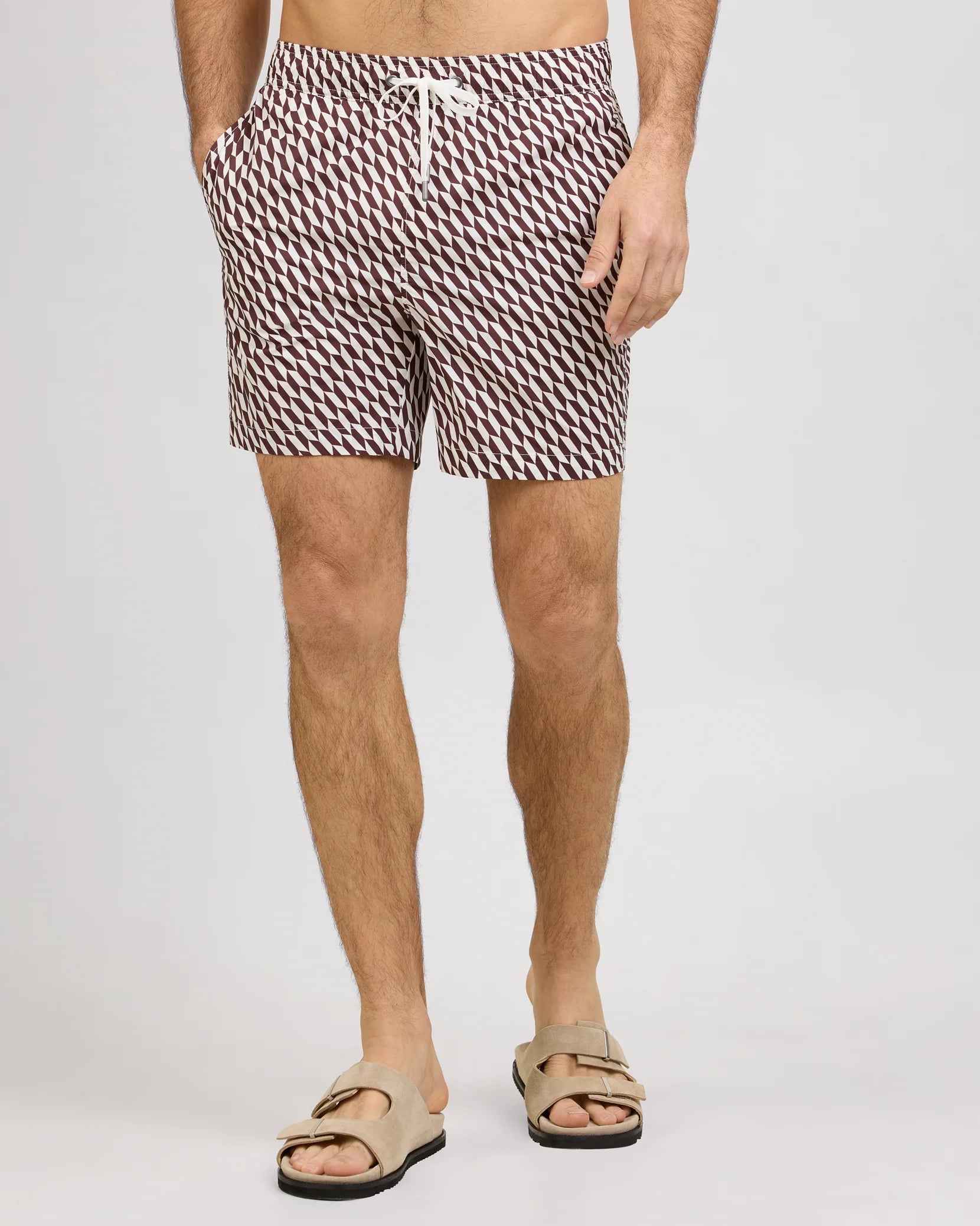 A man stands against a plain light background wearing Onia’s Charles 7″ Swim Trunk, crafted from quick-dry swim fabric with a patterned design and finished with a white drawstring, paired with tan sandals.