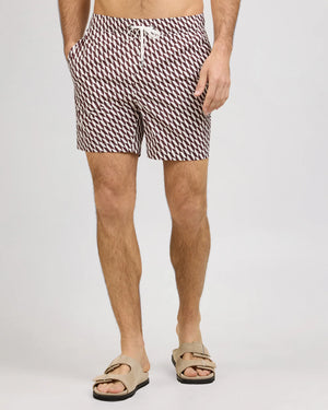 A man stands against a plain light background wearing Onia’s Charles 7″ Swim Trunk, crafted from quick-dry swim fabric with a patterned design and finished with a white drawstring, paired with tan sandals.