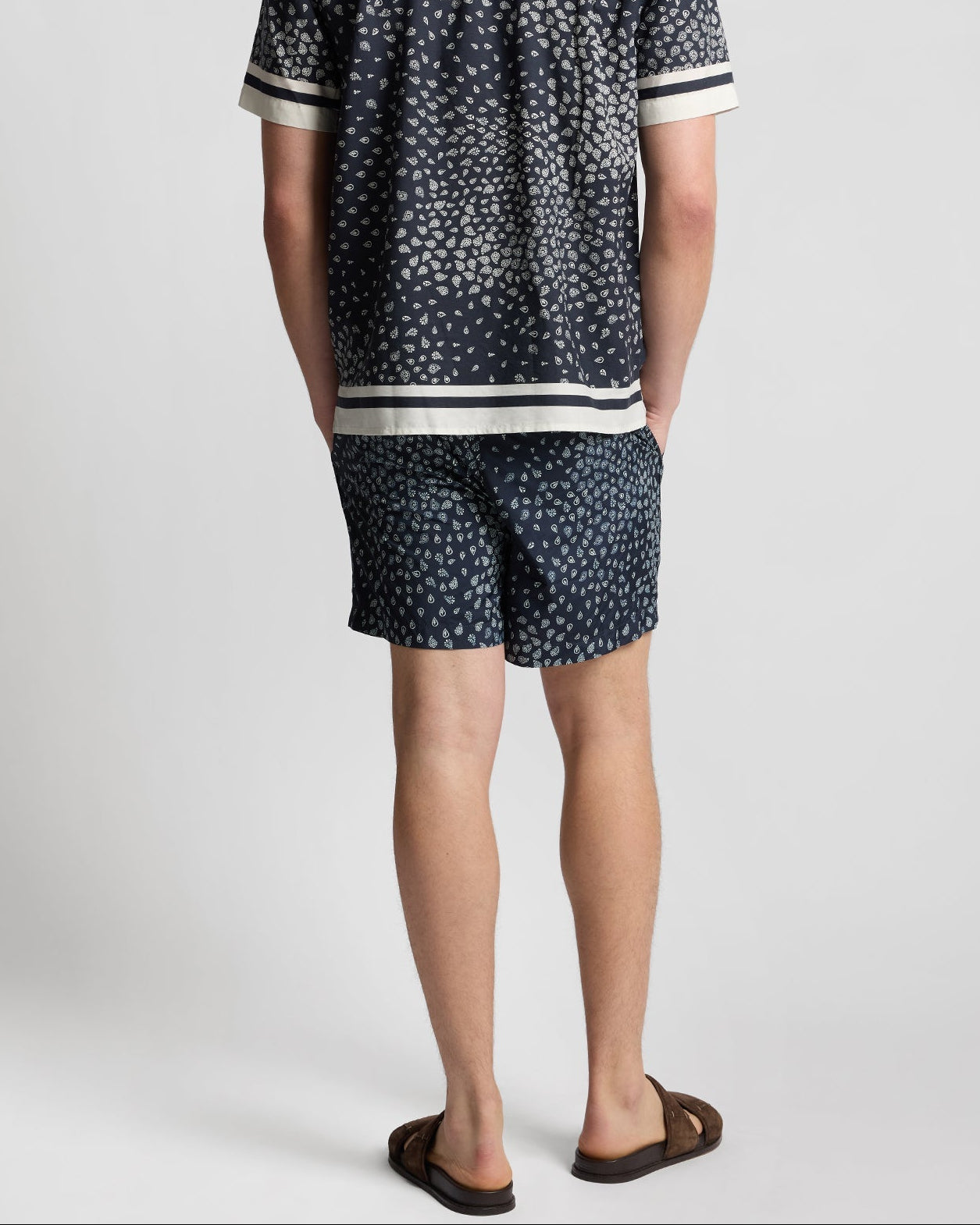Man stands with his back to the camera, wearing a matching navy blue patterned shirt and Onia Calder Buckle 5" Swim Trunk with adjustable waistband, plus brown sandals, against a plain background.