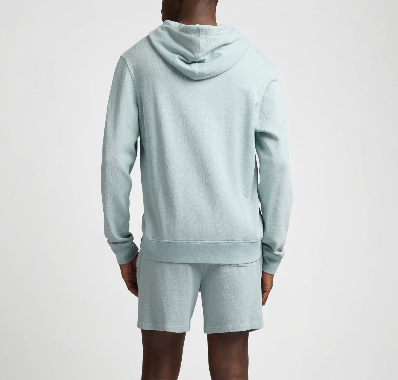 A person stands with their back to the camera, wearing the Onia Terry Pullover Hoodie in light blue and matching heavyweight cotton terry shorts against a plain background.