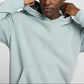 A person adjusts the hood of an Onia Terry Pullover Hoodie in light gray heavyweight cotton terry, shown from head to waist against a neutral background.