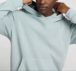 A person adjusts the hood of an Onia Terry Pullover Hoodie in light gray heavyweight cotton terry, shown from head to waist against a neutral background.