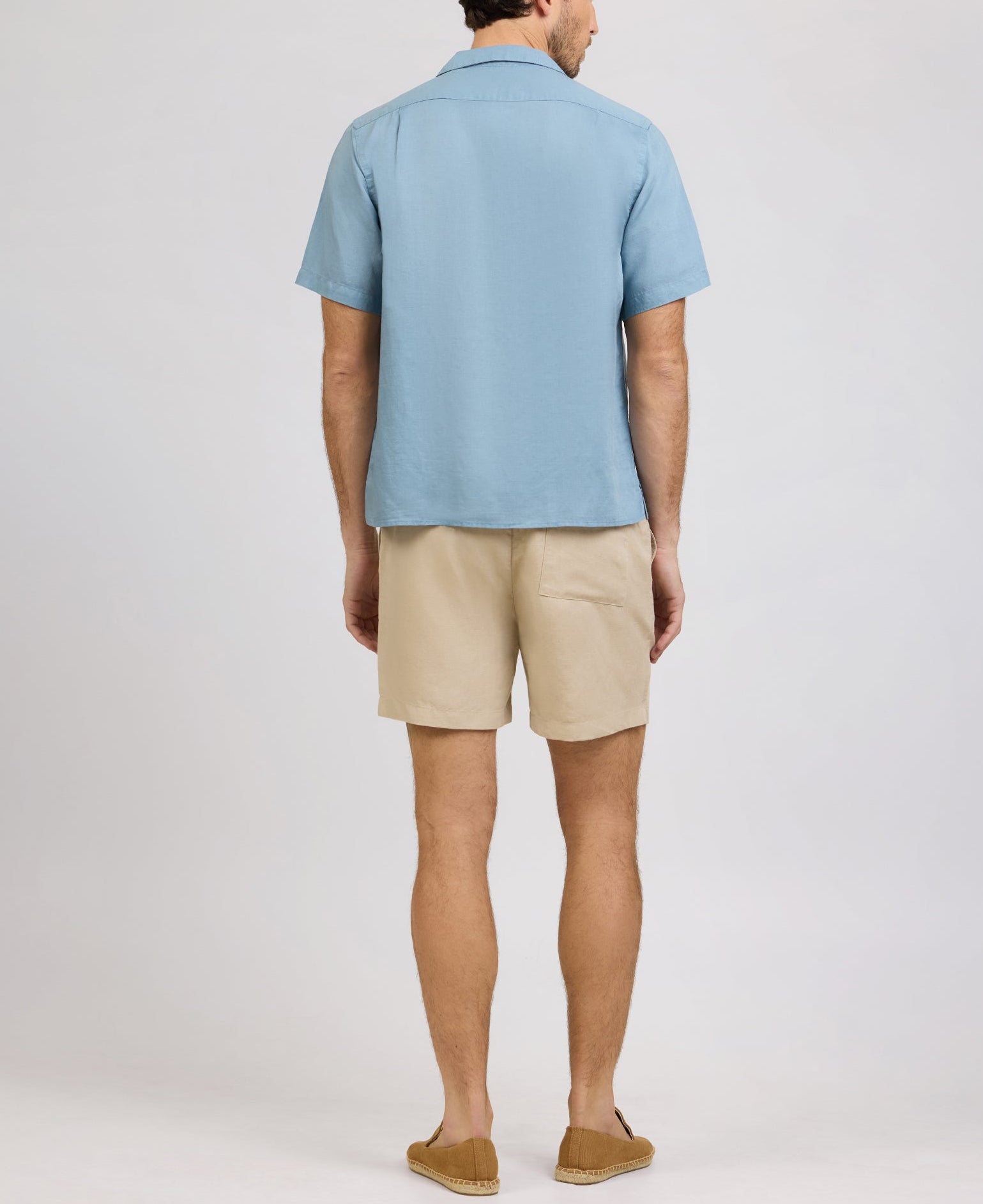 A man faces away, wearing the Onia Air Linen Convertible Vacation Shirt in light blue with short sleeves, paired with beige shorts and brown loafers against a plain light background.