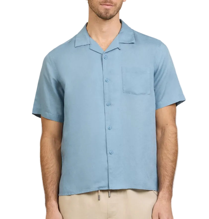 A man wearing the Onia Air Linen Convertible Vacation Shirt in light blue with short sleeves and a chest pocket, paired with beige pants, stands against a white background.