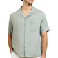 A man wearing the Onia Air Linen Convertible Vacation Shirt in light green with short sleeves and a left chest pocket, paired with white pants, stands with his hands in his pockets.