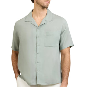 A man wearing the Onia Air Linen Convertible Vacation Shirt in light green with short sleeves and a left chest pocket, paired with white pants, stands with his hands in his pockets.