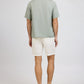 A man faces away from the camera, wearing Onia's Air Linen Convertible Vacation Shirt in light green, paired with white shorts and beige sandals, set against a plain background.