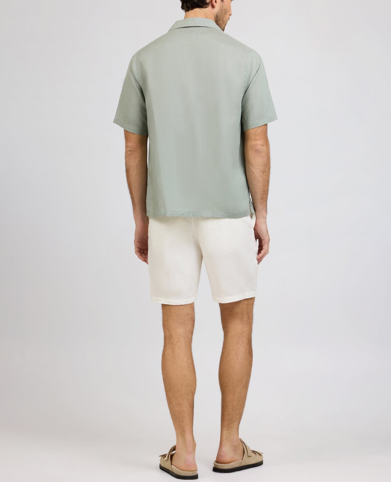 A man faces away from the camera, wearing Onia's Air Linen Convertible Vacation Shirt in light green, paired with white shorts and beige sandals, set against a plain background.