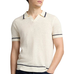 A man wearing the Onia Linen Johnny Collar Polo Sweater—a short-sleeve cream knit with dark trim—stands with one hand in his pocket.