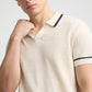 A person models the Onia Linen Johnny Collar Polo Sweater, a cream knit shirt with dark trim and a breathable design, posing with one hand near the Johnny collar.