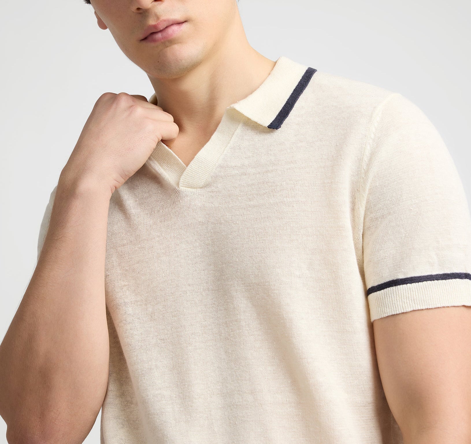 A person models the Onia Linen Johnny Collar Polo Sweater, a cream knit shirt with dark trim and a breathable design, posing with one hand near the Johnny collar.
