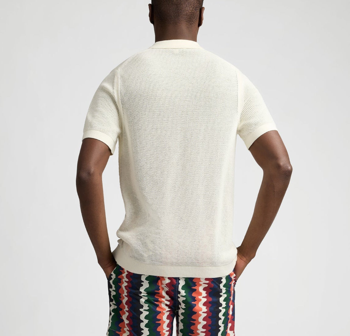A person faces away, wearing the Onia Crochet Knit Button Up, a short-sleeve white shirt, paired with multicolored zigzag shorts against a plain background.