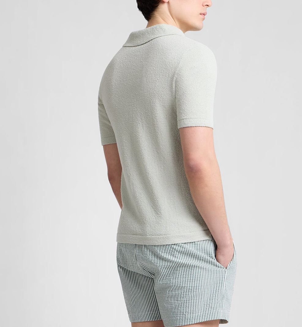 A person stands facing away, wearing the Boucle Knit Johnny Polo by Onia—a light-toned, relaxed fit top—paired with striped shorts, set against a plain background.