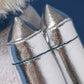 Close-up of two Munro Scottie Dog Space Outfit accessories by Jellycat, featuring metallic silver rocket shapes with textured surfaces, set side by side against a blurred blue and white background for a synthpop feel.
