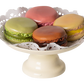 A white pedestal plate holds five hand-painted Maileg Macarons et Chocolat Chaud by Maileg in various colors: pink, green, brown, and tan, with decorative paper underneath them.