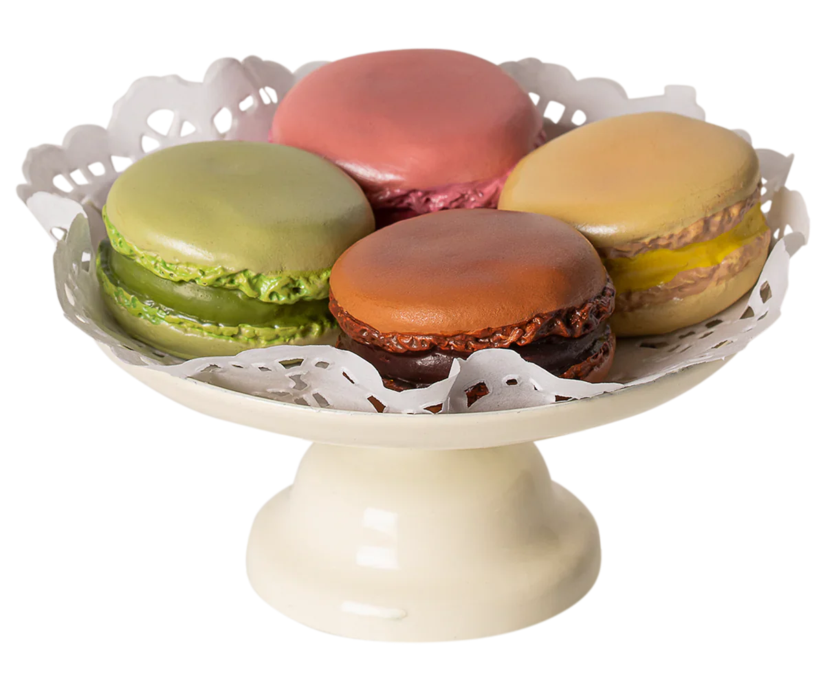 A white pedestal plate holds five hand-painted Maileg Macarons et Chocolat Chaud by Maileg in various colors: pink, green, brown, and tan, with decorative paper underneath them.