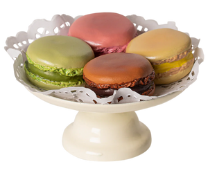 A white pedestal plate holds five hand-painted Maileg Macarons et Chocolat Chaud by Maileg in various colors: pink, green, brown, and tan, with decorative paper underneath them.