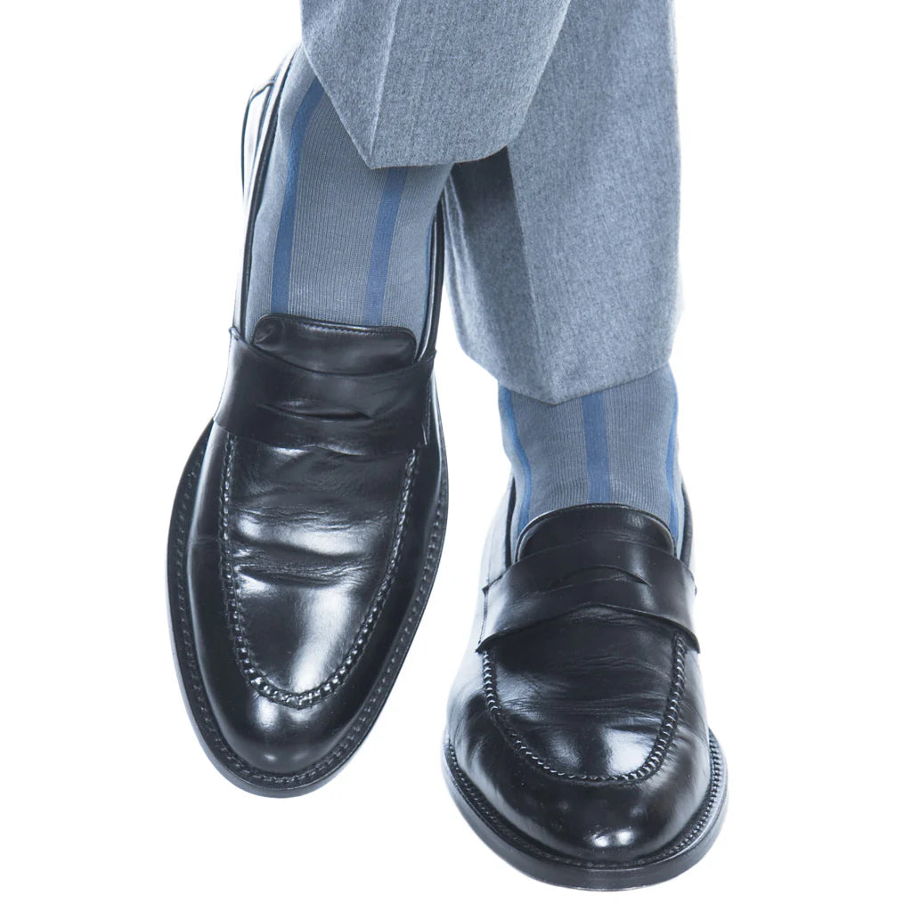 A person wearing gray dress pants, Dapper Classics Steel Gray with Indigo Blue Vertical Stripe Cotton Socks, and black leather loafers with legs crossed at the ankles.