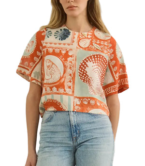 A woman wears a short-sleeved Maeve Top by Hunter Bell NYC in the Lost at Sea print, featuring orange and beige sea-themed designs with a boxy fit, paired with light blue jeans.