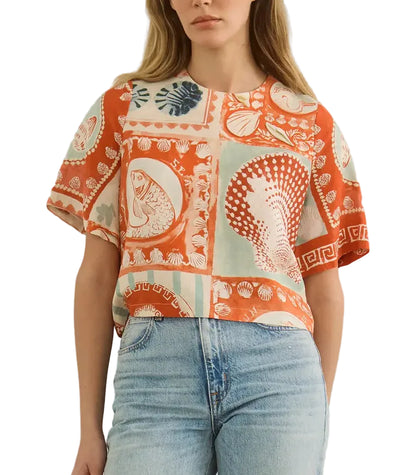A woman wears a short-sleeved Maeve Top by Hunter Bell NYC in the Lost at Sea print, featuring orange and beige sea-themed designs with a boxy fit, paired with light blue jeans.