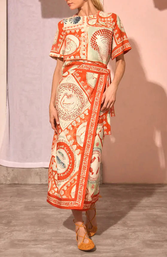 A woman wearing the Maeve Top by Hunter Bell NYC, featuring short sleeves, an orange and cream pattern, a boxy silhouette, and wrap detail, stands on a concrete floor against a neutral background.