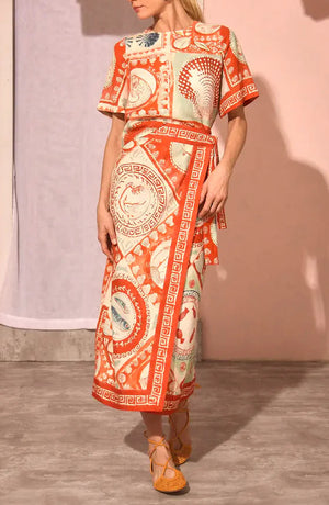 A woman wearing the Maeve Top by Hunter Bell NYC, featuring short sleeves, an orange and cream pattern, a boxy silhouette, and wrap detail, stands on a concrete floor against a neutral background.