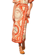 Person wearing an orange and white patterned Paloma Wrap Shirt by Hunter Bell NYC, featuring circular and geometric designs with a waist tie, styled with brown woven shoes.