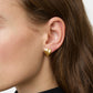 Close-up of a person's ear wearing Lie Studio's Magda Earrings—gold, heart-shaped studs inspired by vintage design; the person has long brown hair and is dressed in black.