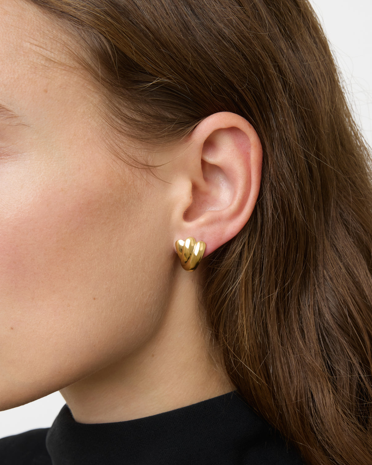 Close-up of a person's ear wearing Lie Studio's Magda Earrings—gold, heart-shaped studs inspired by vintage design; the person has long brown hair and is dressed in black.