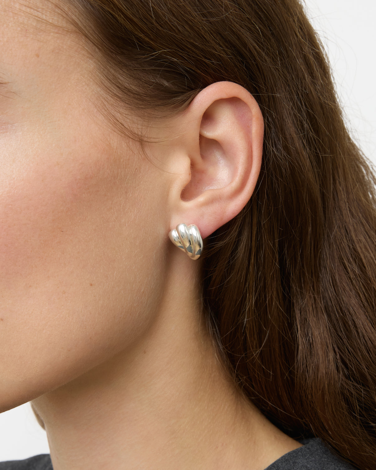 Close-up of a person's ear wearing chunky, sculpted Lie Studio Magda Earrings in sterling silver. The person has long brown hair and a dark top, evoking vintage silhouettes.