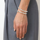 A close-up of a hand wearing two thick 925 sterling silver bangles, including the Lie Studio Maggie Bracelet, resting against gray trousers for a versatile look.