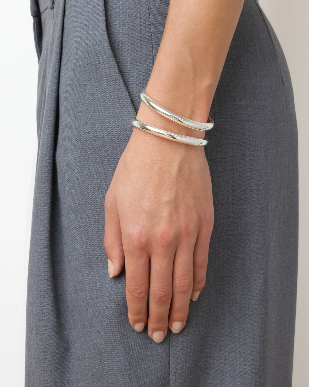 A close-up of a hand wearing two thick 925 sterling silver bangles, including the Lie Studio Maggie Bracelet, resting against gray trousers for a versatile look.