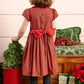 A young girl with brown hair wears the Antoinette Baby Girls' and Girls' Marguerite Red Tartan Smocked Dress, accented with a large red bow, black tights, and red shoes as she stands near Christmas decorations and gifts, facing away.