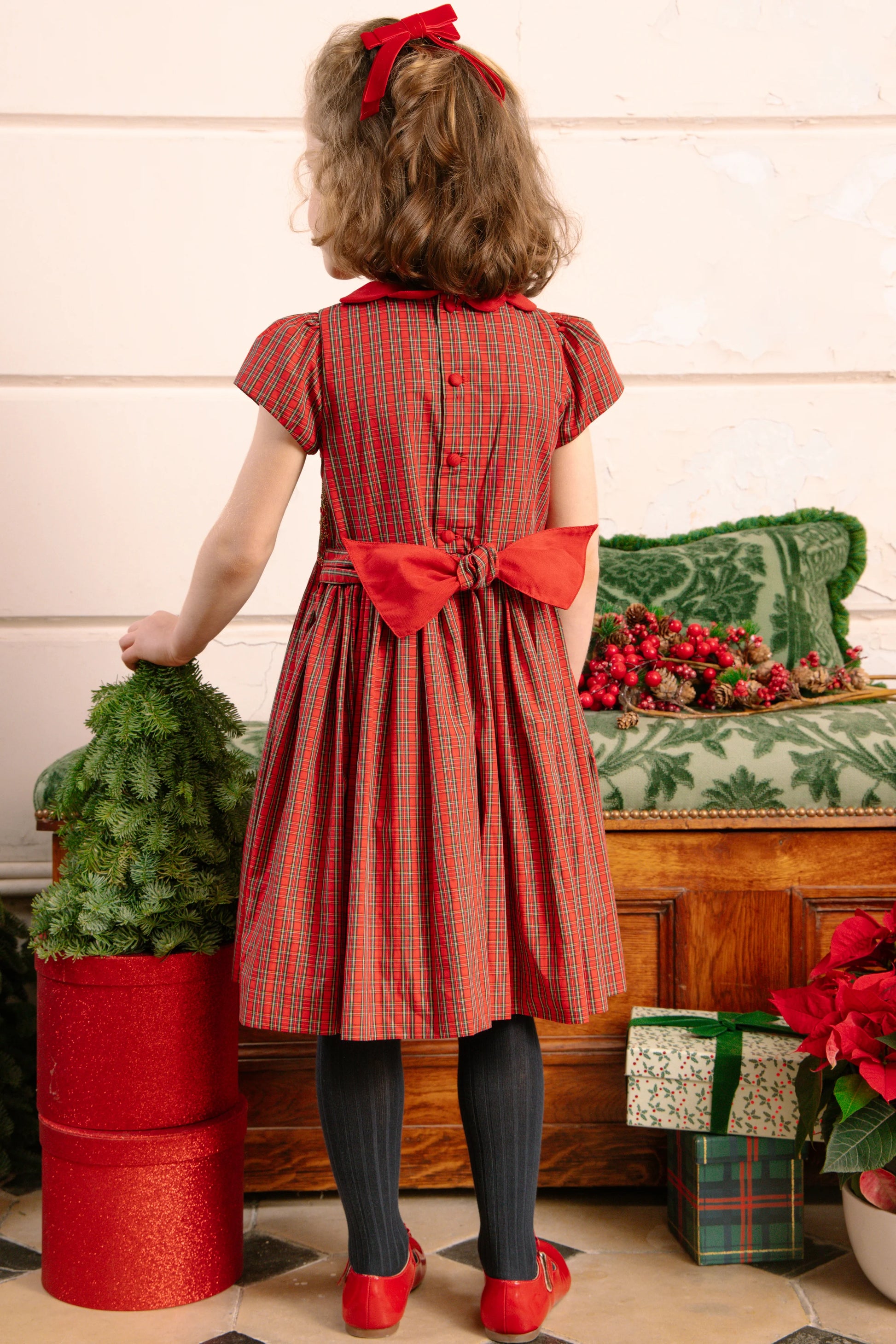 A young girl with brown hair wears the Antoinette Baby Girls' and Girls' Marguerite Red Tartan Smocked Dress, accented with a large red bow, black tights, and red shoes as she stands near Christmas decorations and gifts, facing away.