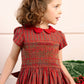 A young girl with curly brown hair wears the Antoinette Baby Girls' and Girls' Marguerite Red Tartan Smocked Dress with a red collar and matching bow, standing indoors and looking to the side—ideal for holiday gatherings.