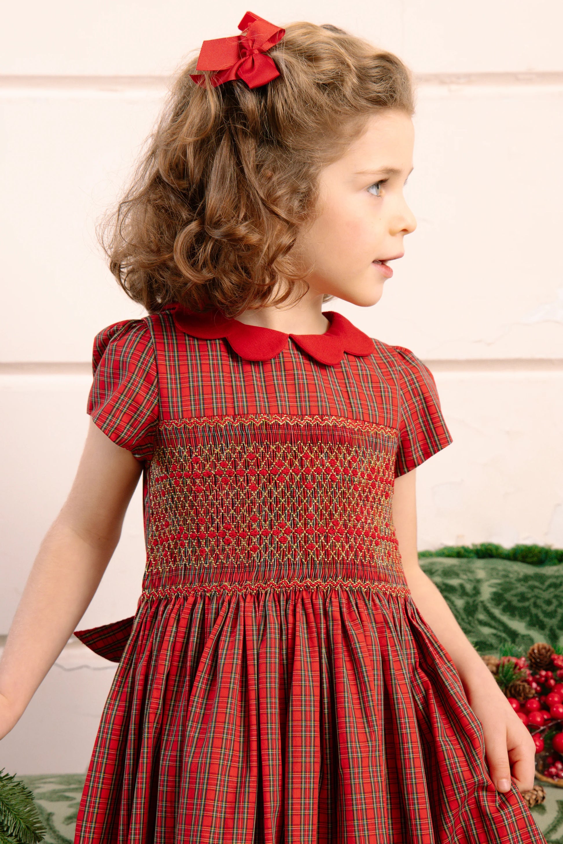 A young girl with curly brown hair wears the Antoinette Baby Girls' and Girls' Marguerite Red Tartan Smocked Dress with a red collar and matching bow, standing indoors and looking to the side—ideal for holiday gatherings.