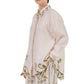 A person wears the Cecilia Shirt by Agua By Agua Bendita, a chic cream button-down with green and yellow floral embroidery on the collar, cuffs, and hem, styled with striped pants.