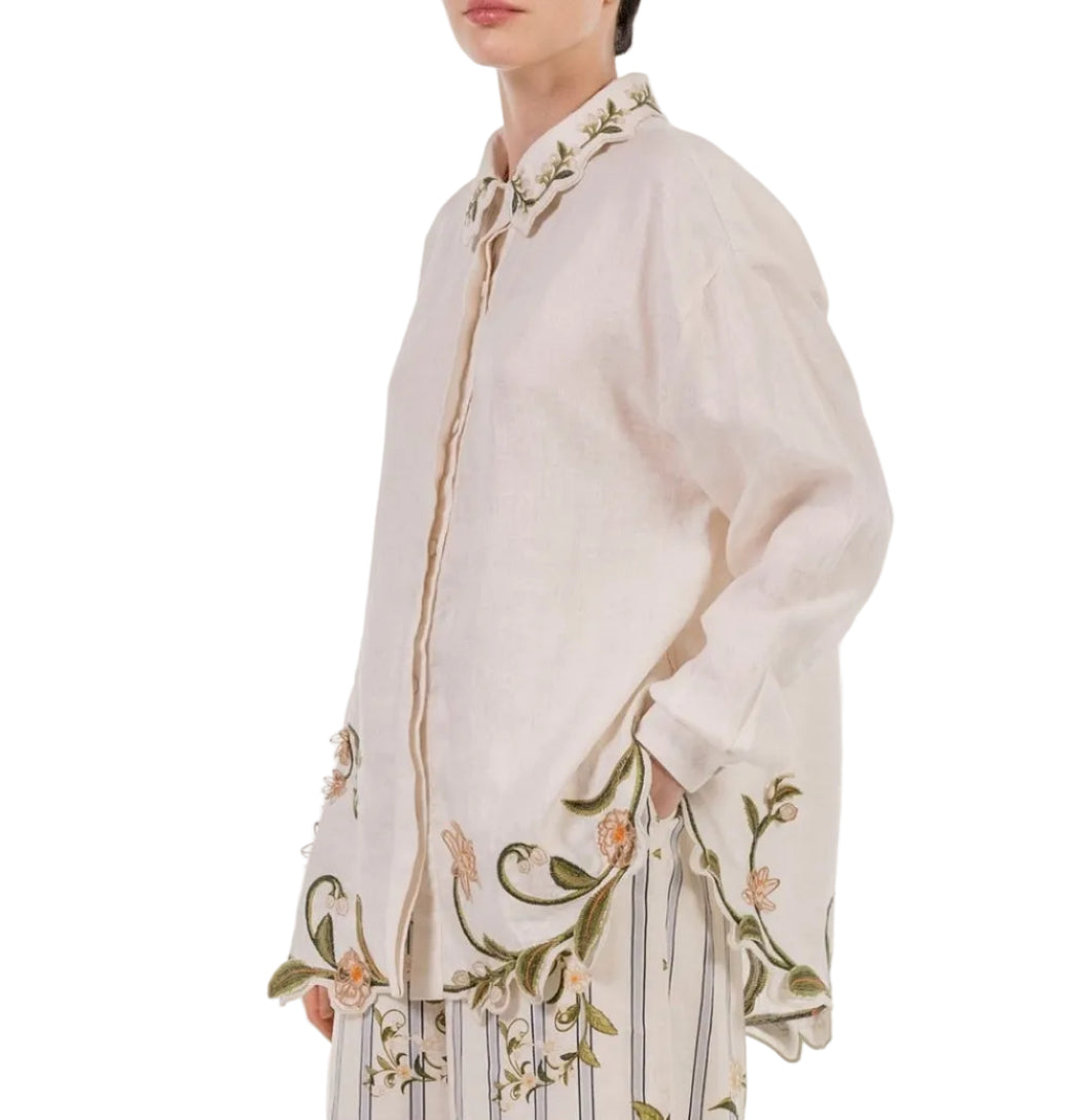 A person wears the Cecilia Shirt by Agua By Agua Bendita, a chic cream button-down with green and yellow floral embroidery on the collar, cuffs, and hem, styled with striped pants.