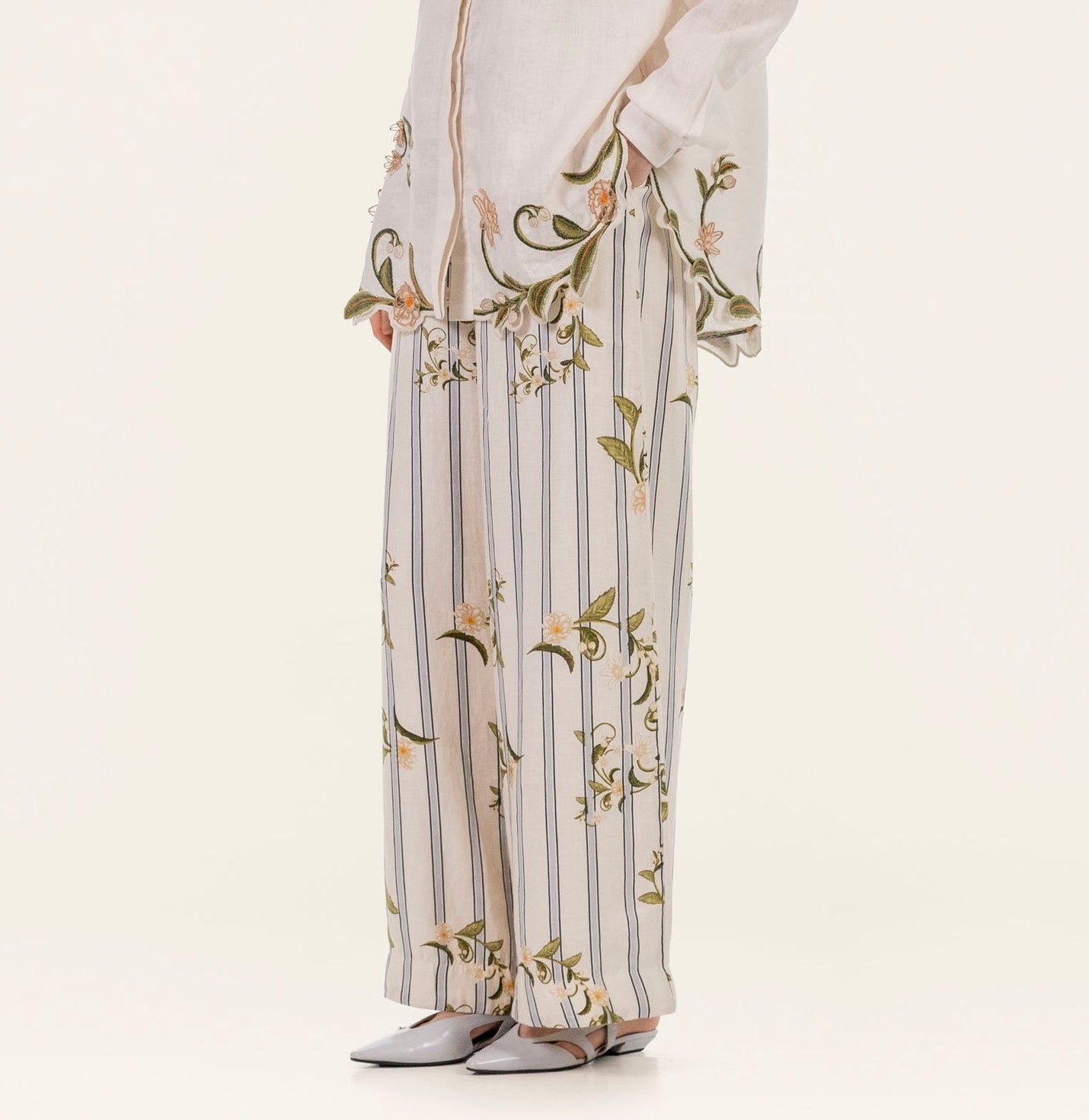Model shown from shoulders down wearing the Tota Pant by Agua By Agua Bendita—linen high-rise floral pants featuring wide-leg stripes and embroidery—with a matching floral-trim top and white pointed-toe shoes, against a plain background.