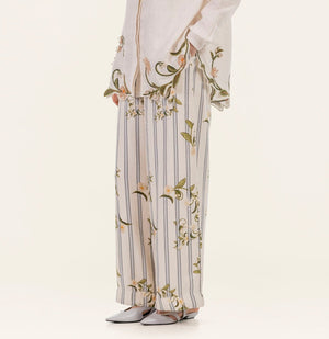 Model shown from shoulders down wearing the Tota Pant by Agua By Agua Bendita—linen high-rise floral pants featuring wide-leg stripes and embroidery—with a matching floral-trim top and white pointed-toe shoes, against a plain background.