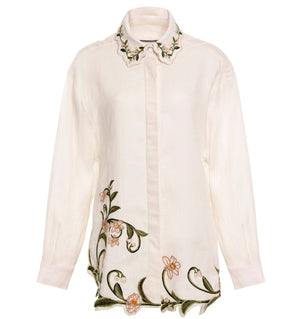 The Cecilia Shirt by Agua By Agua Bendita is a long-sleeve white button-down with floral embroidery in green leaves and orange flowers on the collar and hem. Pair with matching trousers for a polished, coordinated look.