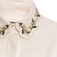 Close-up of the Cecilia Shirt by Agua By Agua Bendita in cream, showcasing a scalloped collar with green and pink floral embroidery—ideal to pair with matching trousers for an elegant ensemble.