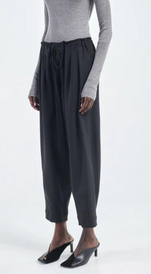 A person wears a grey ribbed long-sleeve top, Maria McManus Barrel Leg Drawstring Trouser, and black heeled mules while standing against a plain light background.