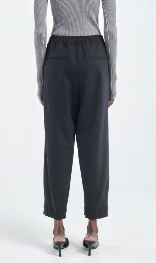 Viewed from behind, person wears Maria McManus Barrel Leg Drawstring Trouser in black Japanese cotton with back pockets, a grey ribbed long-sleeve top, and heeled sandals.