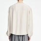 A person with short hair is shown from the back against a plain white background wearing the Maria McManus Pearl Button Blouse in off-white Japanese Naia Renew matte sateen—a loose, long-sleeved style—paired with a gray skirt.