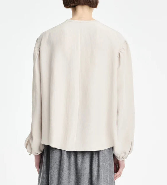 A person with short hair is shown from the back against a plain white background wearing the Maria McManus Pearl Button Blouse in off-white Japanese Naia Renew matte sateen—a loose, long-sleeved style—paired with a gray skirt.